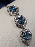 Close-up photo showing the linked floral-shaped silver settings with blue topaz stones and surrounding clear stones.