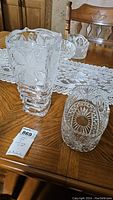 Photo showing clear crystal vase with leaf etching and stepped base alongside rectangular crystal serving dish with detailed cut patterns arranged on wooden table with lace runner.