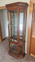 Full view of the curio cabinet showing its wood frame, glass front doors, and lower glass panels. The cabinet has a corner hexagonal shape and stands tall.