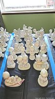 Wide view showing many porcelain Precious Moments figurines arranged on tables, mostly children and angelic characters in soft pastel tones.