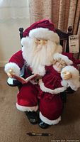 Front view of Santa Claus figurine seated in wooden chair holding open book and child figure, glasses with one lens missing.