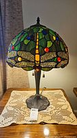 Full view of Tiffany style lamp shown on a wooden table with lace cloth underneath. The stained glass shade has dragonfly motif with colorful glass pieces and the metal base has ornate floral design.