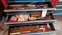 Drawer view showing multiple hand tools including files, chisels, a tape measure, and paint brushes inside red tool chest drawers.