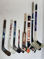 Eight assorted hockey sticks, mix of souvenir and playing sticks, different brands, colors, and sizes laid flat
