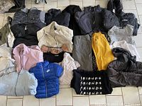 Assorted men's, women's, and kids clothing items arranged on floor including jackets, pants, and shirts of various colors and styles.