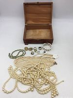 Open wooden jewelry box with assorted fashion jewelry laid out in front: multiple pearl-like bead necklaces, white geometric bracelet, metal clip-on earrings, and gold-tone braided bracelet.