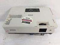 Top front view of white Epson projector with lens, focus dial, and security sticker visible.