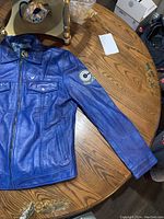 Blue leather jacket laid out flat showing front and sleeve Capsule Corp logo.