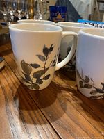 Four white ceramic coffee mugs with gray and muted green floral designs on the side shown clustered together on wooden surface.