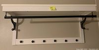 White wall-mounted wooden shelf with black metal hanging rod held by black metal brackets, underneath are five black wooden pegs.
