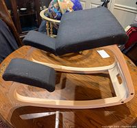 Angle view of ergonomic kneeling chair showing black fabric cushions and light wooden frame, placed on wood surface.
