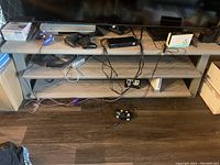 Front view of the wooden media stand with three shelves. Cables, game controllers, and electronic devices visible on and around the stand, with TV on top.