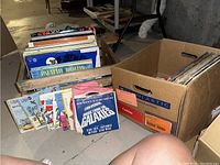 Photo of two boxes containing numerous vinyl records, both albums and 45s, on a basement floor with diverse genres visible