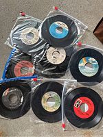 Multiple black vinyl 45 RPM records with colorful labels in plastic bags, featuring artists Eagles, Queen, The Playmates, The Righteous Brothers, Drifters and more.