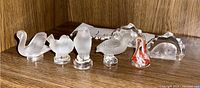 Six signed crystal animal figurines shown on wooden shelf, including four clear/frosted birds, one red white swirl figurine, and one arch-shaped animal figurine.