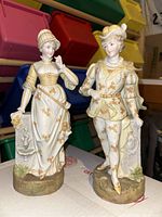Front view of two ceramic figurines, a man and a woman in ornate historical costumes with floral gold detailing.
