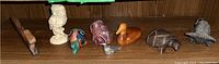 Nine animal figurines displayed on a wooden shelf: carved wood turtle, metal bull, wood carved squirrel and duck, metal bird bell, two painted pottery figurines, and composite owl figurine.