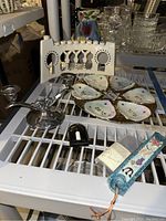 Overview of all Judaica items on a white metal shelf: ceramic menorah, seder plate, metal candle holder, plastic plug-in light, decorative mezuzah with scroll.
