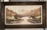 Front view of the framed painting depicting a river or lake with two houses and rocky hills, signed by artist Ann.
