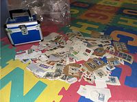 Photo of multiple loose postage stamps and small stamp envelopes scattered on a colorful foam play mat with the blue metal case partially visible.