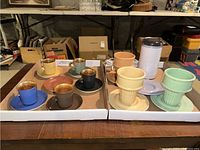 Overview of colorful Figgjo Flint Norway tea cups and saucers along with pastel-colored dessert cups and a Cuisinart coffee grinder on a table in a basement/storage room.