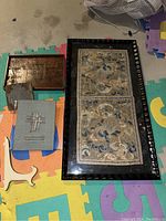 Three framed Asian style tapestry wall art pieces displayed on a foam floor mat, including two large embroidered tapestries in black wood frames and one smaller acrylic framed relief art piece.