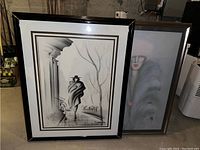 Two framed artworks leaning against a wall in a dimly lit setting: one black and white drawing showing a cloaked figure walking by columns and stairs, the other a stylized portrait with pale face and pink lips.