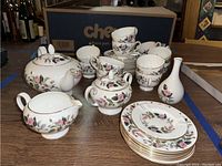 Photo showing complete set arranged on table: teapot, sugar bowl, creamer, teacups, saucers, side plates, bud vase, and ashtray all with floral rose and gold trim design.