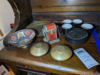Wide shot showing entire lot items on wooden surface including bowl, brass dishes, pencil sharpener, black espresso cups and saucers.