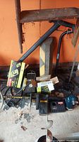 Overview of lot showing chain saw, Ryobi charger box, handheld drill, and other accessories on garage floor under a wooden shelf
