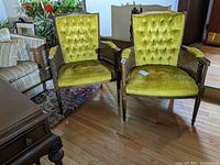 Two vintage wooden armchairs with green velveteen upholstery and caned armrests, shown from front view in a room with hardwood floor.