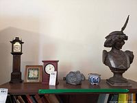 Overview photo showing all items on table: two tall wooden clocks, gold frame desk clock, red desk clock, gray rabbit figurine, blue and white planter, and Renaissance bust sculpture.
