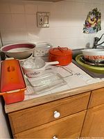 Countertop view showing the orange enameled cast iron pot with lid, Pyrex Friendship pattern Cinderella bowl, CorningWare Blue Cornflower rectangular casserole dish, red-orange glass loaf baking pan, and some wooden bowls.
