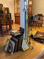 Photo showing a collection of various empty picture frames stacked on the floor in different sizes and finishes, along with some round wall plates and a tall decorative metal wall plate holder leaning against a blue wall.