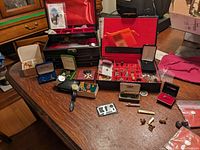Overview of lot items laid out on table including jewelry boxes, tie clips, cufflinks, ring boxes, watches, collector pins, and charms