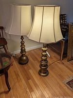 Photo of two brass finish table lamps standing on wood floor showing full lamp with beige fabric shades and cords.