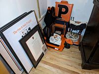 Photo showing a corner with various Princeton items: a large foam orange 'P' sign, orange and black backpacks, folded white t-shirt, a small tiger plush doll on a chair, coasters, a framed certificate, and wall art leaning against the wall.