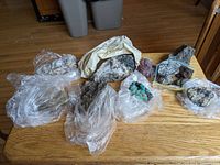 Photo showing a group of roughly 10 rocks, mineral samples and stones spread out on a wooden table, wrapped mostly in plastic bags or plastic wrap.