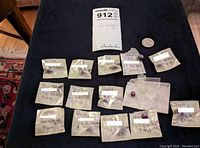 Collection of loose ruby gemstones in small taped bags laid out on a black surface with a quarter for scale and a MaxSold tag labeled 912.