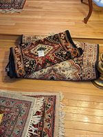 Three oriental rugs are shown folded on a wood floor, revealing intricate colorful pattern and fringed edges.