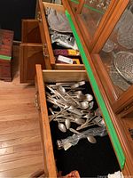 Open wooden drawers showing a mixed collection of silver plated flatware including forks, spoons, and knives, resting on black liners.