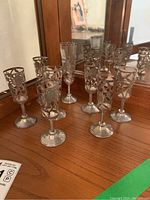 Group of nine sterling silver cordial holders arranged on wooden surface in front of mirror, showing decorative filigree patterns and bases.