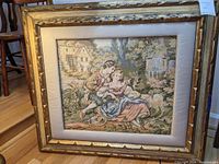 Framed tapestry artwork showing a romantic couple in an outdoor garden setting with architectural background, in a decorative frame.
