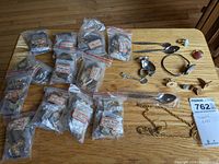 A wooden table displaying multiple small clear plastic bags containing Tiger Eye pendants with labeled quantities, loose chains, metal spoons, and assorted costume jewelry items arranged around the bags.