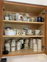 Wooden cabinet shelves holding multiple ceramic coffee mugs and teacups arranged in groups, some vintage with unique patterns.