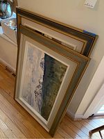 Two framed paintings leaning against wall; one gray-and-gold frame in front, one white frame behind