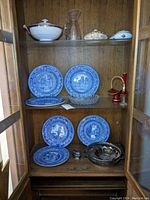 Cabinet display with Wedgwood Princeton blue transferware plates on the middle and bottom shelves, porcelain serving bowl and covered dishes on the top shelf, along with ruby glass and glassware decor items