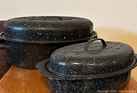 Two black speckled roasting pans with lids on a wooden surface against a plain wall.