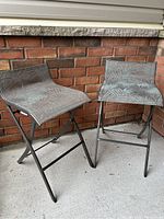 Two foldable stools with dark metal frames and leaf-patterned fabric seats placed against a brick wall on a concrete floor.