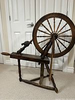 Side view of antique wooden spinning wheel showing round wheel with multiple spokes and foot pedal lever.